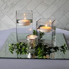 candles are lit in glass vases with greenery on a table top, surrounded by marble tiles