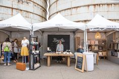 an outdoor market with people shopping and selling items under white tented umbrellas, in front of large industrial silos