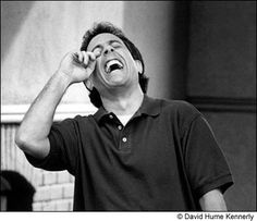 black and white photograph of a man laughing with his hands on his head while standing in front of a brick wall