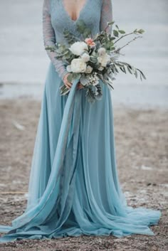 a woman in a long blue dress holding a bouquet and standing next to the water