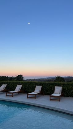 lounge chairs line the edge of a swimming pool at dusk with an moon in the distance