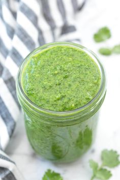 a glass jar filled with green sauce on top of a white table cloth next to cilantro leaves