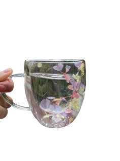 a hand holding a glass cup with flowers on it and water in the bottom, against a white background