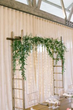 a wooden table topped with white chairs under a window covered in sheer curtains and greenery