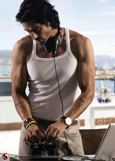 a man in white tank top standing next to a table with a laptop and headphones on