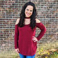 a woman standing in front of a brick wall wearing blue jeans and a red sweater