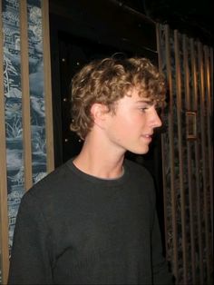 a young man with curly hair standing in front of a metal gate and looking off into the distance