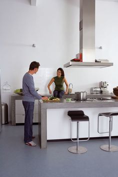 two people in a kitchen preparing food on the counter and stools next to them