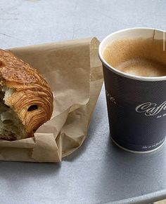 a cup of coffee sitting next to a pastry on top of a table near a paper bag