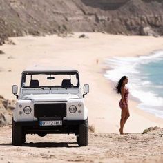 a woman standing on the beach next to a white jeep parked in front of an ocean