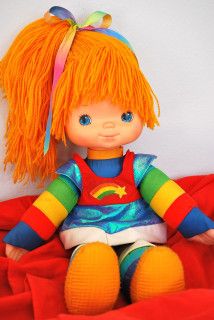 a small doll with orange hair sitting on a red bed sheet and looking at the camera