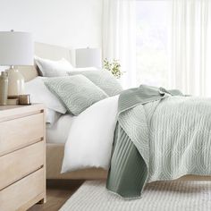 a bed with white sheets and pillows in a bedroom next to a dresser, lamp and window