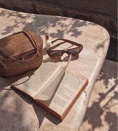 an open book, sunglasses and purse sitting on top of a cement table outdoors next to a tree