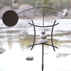 a weathervane with three stones balanced on it's stand in front of a body of water