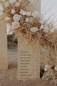 a wooden sign with flowers on it sitting in the sand next to some dry grass