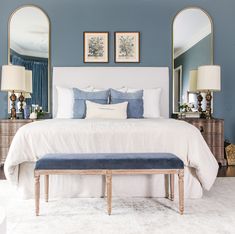 a bedroom with blue walls, white bedding and two mirrors on the wall above it