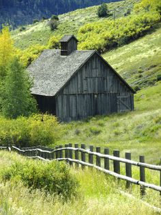 an old barn sits in the middle of a grassy field with trees and mountains behind it