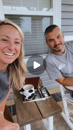 a man and woman sitting at a wooden table with coffee in front of them on the porch