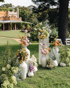 several white vases with flowers in them sitting on the grass near some trees and bushes