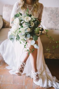 a woman in white dress holding a bouquet of flowers and greenery on her feet