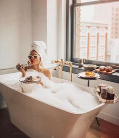 a woman sitting in a bathtub with a towel on her head and holding a drink