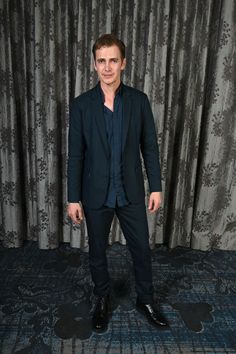 a man standing in front of a curtain with his hands in his pockets and wearing a black suit