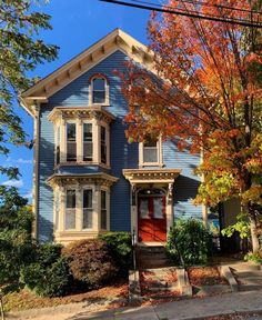 a blue two story house with red door and trees in the front yard on a fall day