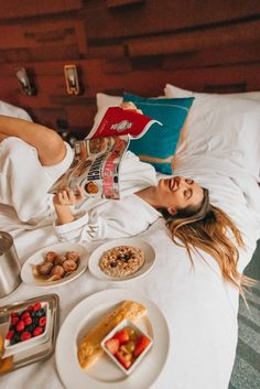 a woman laying in bed reading a book and eating breakfast on the bed with other foods