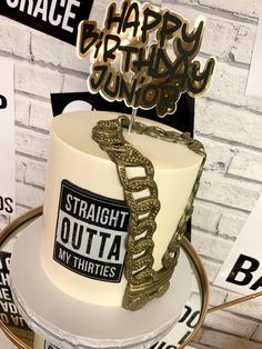 a white and gold birthday cake sitting on top of a metal stand in front of a brick wall