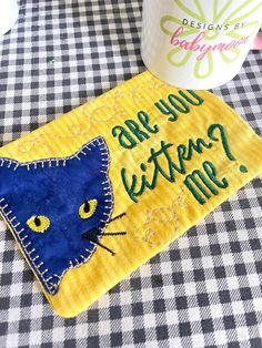 there is a blue and yellow cat on the table next to a coffee cup that says, are you kittens me?