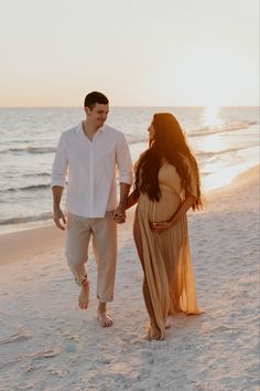 a man and woman holding hands walking on the beach at sunset, with the ocean in the background