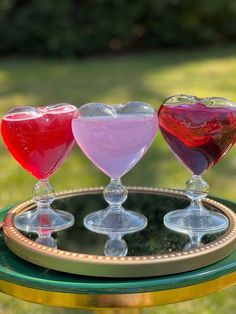 three heart shaped glasses sitting on top of a glass plate in the grass, with one filled with liquid