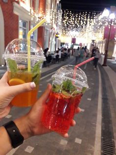 two people holding up drinks in their hands on the street with lights strung above them