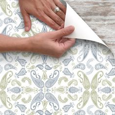 two hands on top of a piece of paper next to a white wallpaper with blue and green paisley designs