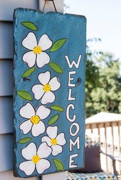 a welcome sign hanging from the side of a house with white flowers painted on it