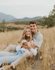 a man and woman sitting on the ground in tall grass with mountains in the background