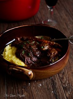 a wooden bowl filled with meat and mashed potatoes next to a glass of wine