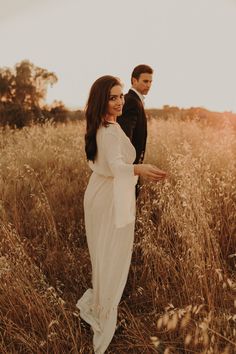 a pregnant woman standing in tall grass next to a man wearing a tuxedo