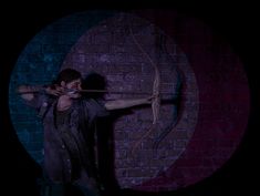 a man holding a bow and arrow in front of a brick wall with a circle behind him