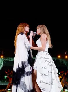 two women in white dresses standing next to each other on stage with one holding a microphone