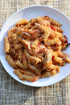 a white bowl filled with pasta and sauce on top of a woven table cloth next to a wooden spoon