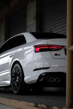 the rear end of a white car parked in front of a garage door with its lights on