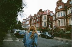 a woman is walking down the street in front of some houses with cars parked on both sides