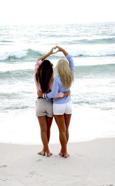 two women standing on the beach with their arms around each other making a heart shape