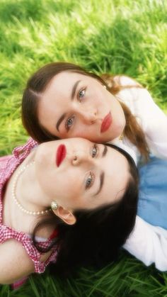 two women laying in the grass with their heads on each other's shoulders and looking at the camera