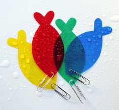 three colorful fish shaped paper clips on a white surface with drops of water around them