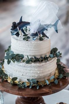 a three tiered cake with white frosting and greenery on top is topped with an origami fish decoration