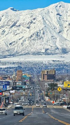 cars are driving down the street in front of snow covered mountains and buildings on either side