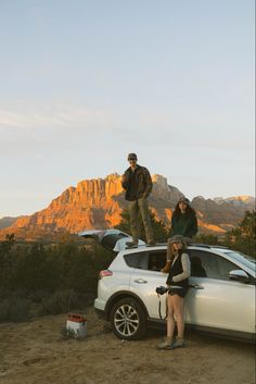 three people standing on the roof of a white car in front of mountains and trees