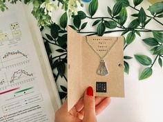 a person holding up a book with a necklace on it next to a plant and a sheet of paper